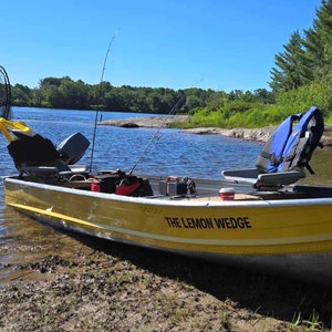 May include: A yellow aluminium fishing boat named THE LEMON WEDGE with two grey swivel seats, a blue life vest, two fishing rods, and a black motor. The boat is resting on a muddy shoreline next to a body of water under a clear blue sky.