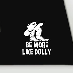 May include: A black surface featuring a white graphic of a cowboy hat resting on a pair of cowboy boots decorated with small stars and hearts. Below the graphic, the text reads BE MORE LIKE DOLLY in a bold, white, sans-serif capital font.