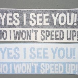 May include: Two rectangular bumper stickers with white text on a dark background and white text on a light blue background. Both stickers feature the text: YES I SEE YOU! NO I WON'T SPEED UP! The lettering is in a bold, sans-serif font style.