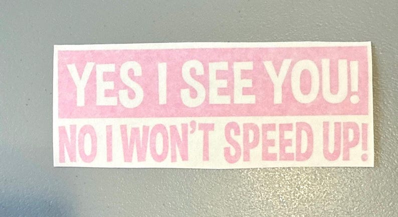 May include: A rectangular sticker with a light pink background and white text that reads YES I SEE YOU! NO I WON'T SPEED UP! The lettering is in a bold, sans-serif font, arranged in two lines with an exclamation point at the end of each.