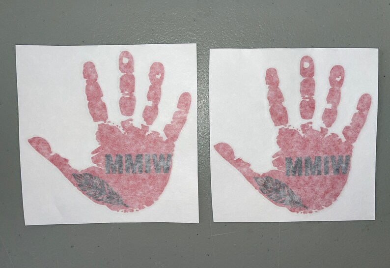 K&ouml;nnte beinhalten: Zwei rote Handabdruck-Aufkleber mit einer schwarzen Feder und dem Text "MMIW" auf wei&szlig;em Hintergrund.