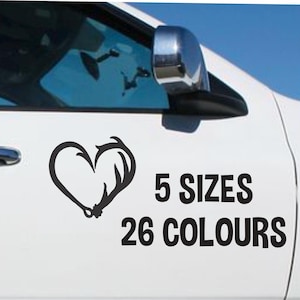 May include: A black vinyl decal applied to a white car door, featuring a heart shape formed by a fishing hook and an antler. The text to the right of the graphic reads 5 SIZES 26 COLOURS in a bold, black, sans-serif font, indicating available options.