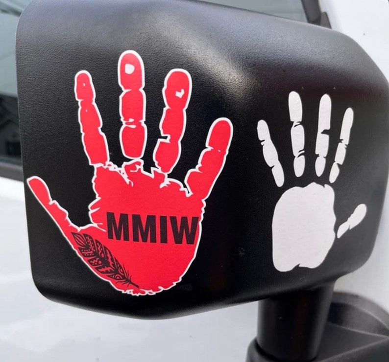 K&ouml;nnte beinhalten: Ein schwarzer Autospiegel mit zwei Handabdruck-Aufklebern. Der linke Handabdruck ist rot mit dem Text "MMIW" und einem Federdesign. Der rechte Handabdruck ist wei&szlig;.