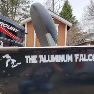 May include: A black aluminium boat with a Mercury outboard motor. The side of the boat features a white graphic of the Great Lakes and the text THE ALUMINUM FALCON in white lettering. A grey seat is visible inside the boat near the motor.
