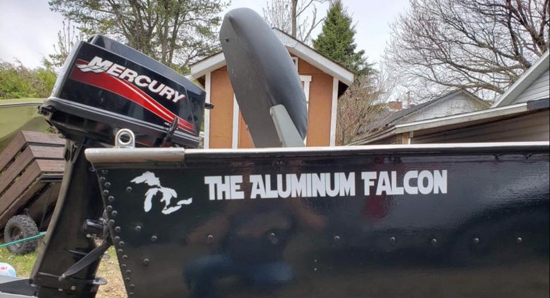 May include: A black aluminium boat with a Mercury outboard motor. The side of the boat features a white graphic of the Great Lakes and the text THE ALUMINUM FALCON in white lettering. A grey seat is visible inside the boat near the motor.