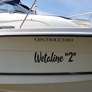 May include: A white boat hull featuring black lettering that reads ON3842300 and the name Wetaline "2" in a cursive script. The vessel includes a metal railing along the top edge and a dark blue canopy visible at the very top of the frame.