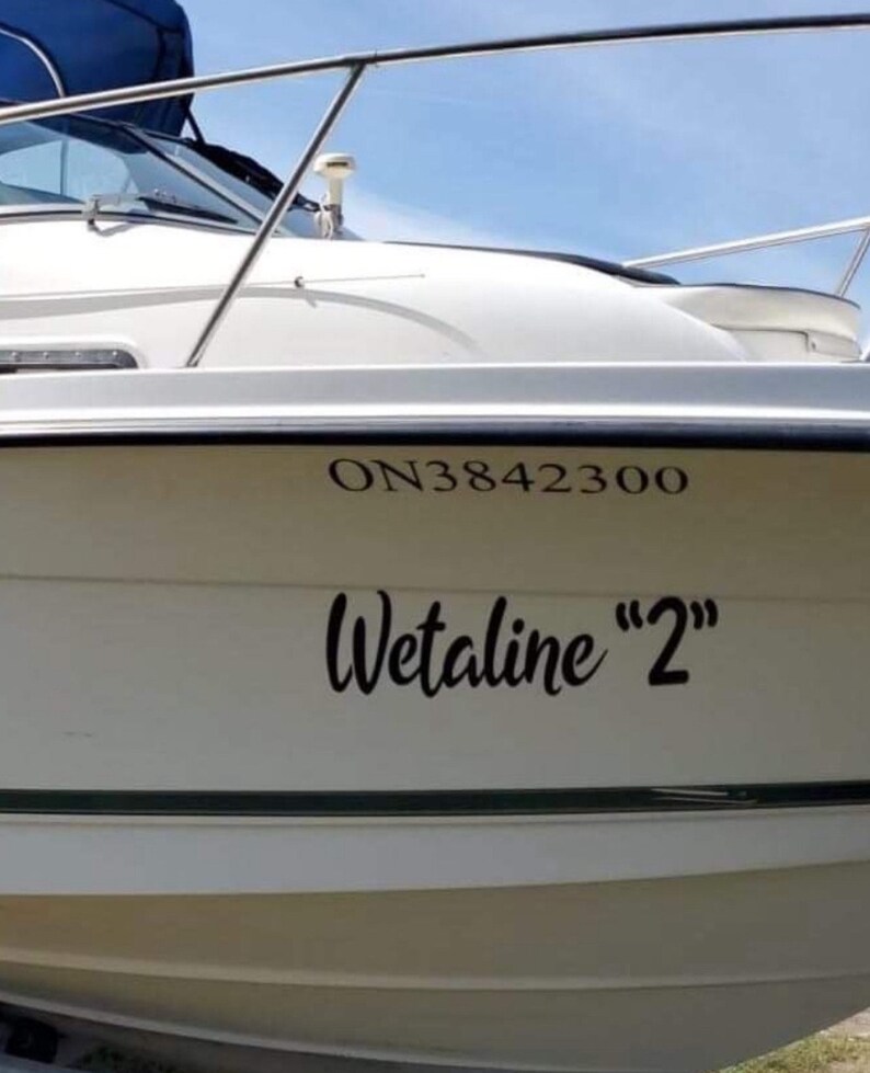May include: A white boat hull featuring black lettering that reads ON3842300 and the name Wetaline "2" in a cursive script. The vessel includes a metal railing along the top edge and a dark blue canopy visible at the very top of the frame.
