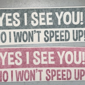 May include: Two rectangular bumper stickers with white text on colored backgrounds. The top sticker is dark gray with white text, and the bottom sticker is pink with white text. Both read: YES I SEE YOU! NO I WON'T SPEED UP!