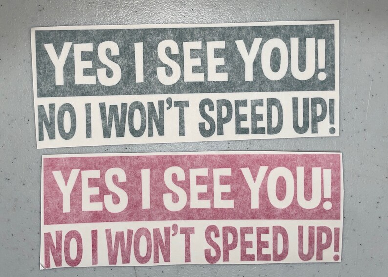 May include: Two rectangular bumper stickers with white text on colored backgrounds. The top sticker is dark gray with white text, and the bottom sticker is pink with white text. Both read: YES I SEE YOU! NO I WON'T SPEED UP!