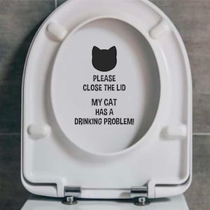 Op de afbeelding: Een witte toiletbril met een zwart katten silhouet en de tekst "PLEASE CLOSE THE LID, MY CAT HAS A DRINKING PROBLEM!". De bril is gesloten en de tekst staat in het midden van de deksel.