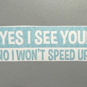 May include: A rectangular bumper sticker with a light blue textured background and white bold sans-serif text that reads YES I SEE YOU! NO I WON'T SPEED UP! The sticker has a clean white border around the entire rectangular design.
