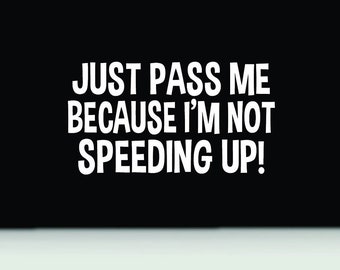 Op de afbeelding: Een zwart rechthoekig bord met dikke witte hoofdletters die luiden: JUST PASS ME BECAUSE I'M NOT SPEEDING UP! De tekst is gecentreerd op de zwarte achtergrond, met een dunne witte horizontale lijn die de onderrand scheidt.
