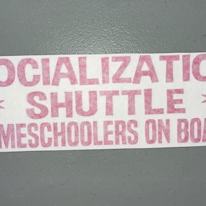 May include: A rectangular white bumper sticker featuring bold, pink, block-style lettering that reads SOCIALIZATION SHUTTLE HOMESCHOOLERS ON BOARD. The text is flanked by two pink, eight-pointed star symbols, one on each side of the word SHUTTLE.
