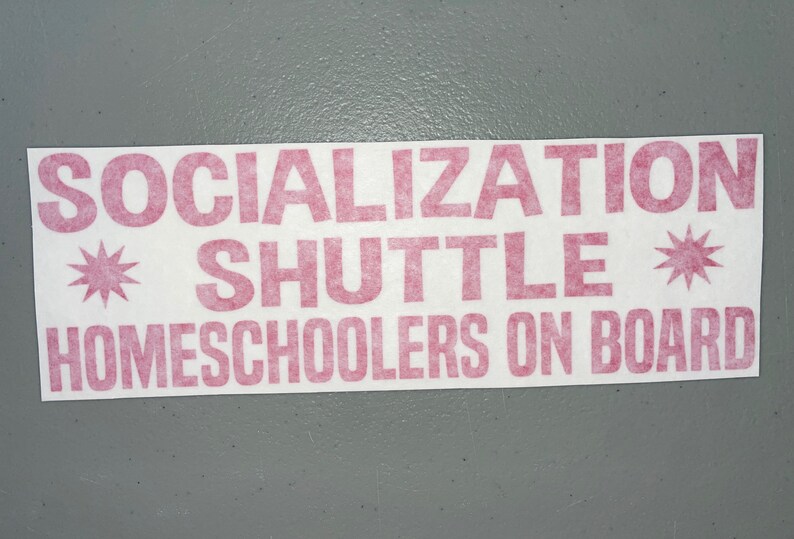 May include: A rectangular white bumper sticker featuring bold, pink, block-style lettering that reads SOCIALIZATION SHUTTLE HOMESCHOOLERS ON BOARD. The text is flanked by two pink, eight-pointed star symbols, one on each side of the word SHUTTLE.