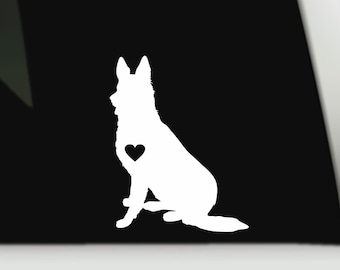 May include: A white vinyl decal featuring the silhouette of a German Shepherd dog sitting in profile. The dog's chest area contains a small, solid black heart cutout, creating a contrast against the white body of the canine figure.