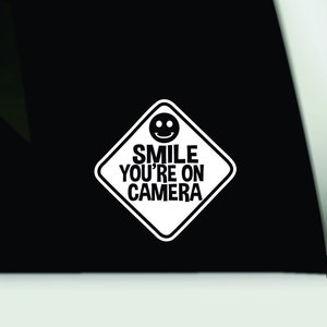 以下が含まれることがあります： 白いひし形のステッカーで、笑顔の顔のアイコンと「SMILE YOU'RE ON CAMERA」という黒い文字が特徴です。窓やその他の表面に貼るように設計されています。
