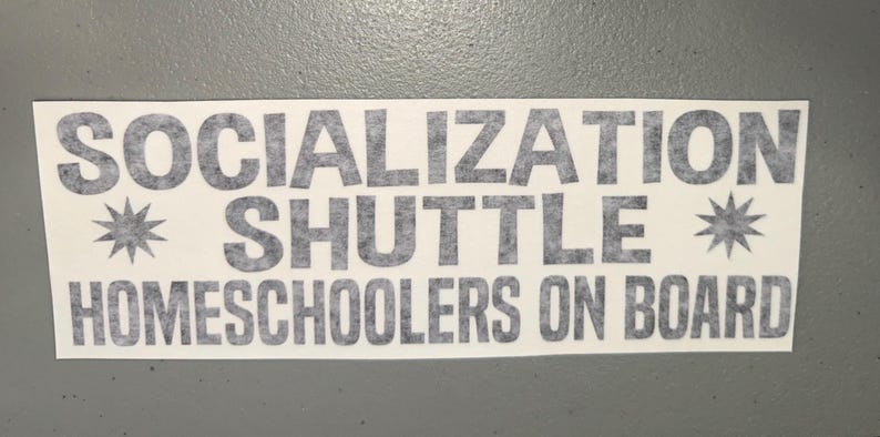 May include: A rectangular white bumper sticker featuring the dark gray text SOCIALIZATION SHUTTLE HOMESCHOOLERS ON BOARD in a bold sans-serif font. The text is arranged in three lines with a small eight-pointed star icon on each side of the middle line.