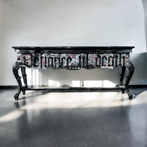 May include: Black console table with ornate legs and a lower shelf. The table's front features the text "Defiance til death" in a gothic font, with a collage of images beneath the text. The table is set against a white wall.
