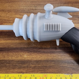 May include: A gray and black plastic toy ray gun with a long barrel and a handle. The gun has a textured surface and a small, round knob on top.