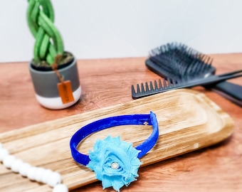 Puede incluir: Una diadema de terciopelo azul con una flor de tela azul claro y un strass en el centro.