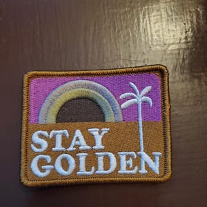 May include: Embroidered patch with a rainbow, palm tree, and the words "STAY GOLDEN" in white block letters. The patch has a brown and purple background with a gold border. The patch is rectangular.