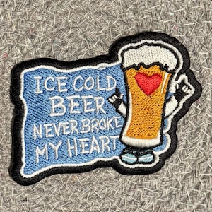 May include: Embroidered patch featuring a cartoon beer glass with a red heart, the text "ICE COLD BEER NEVER BROKE MY HEART" on a blue background, and a black border. The beer glass has a white foamy top.
