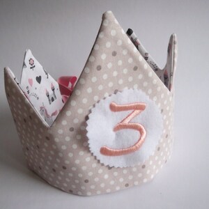 Peut inclure: Une couronne en tissu beige à pois blancs et un chiffre "3" en feutre rose sur un cercle blanc. La couronne est décorée d'un ruban rose et d'un tissu blanc à motifs roses, noirs et blancs.