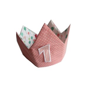 Può includere: Una corona di tessuto rosa con pois bianchi e un numero "1" bianco cucito sul davanti. La corona è realizzata in tessuto ed è progettata per il compleanno di un bambino.