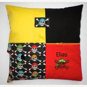 Könnte beinhalten: Ein schwarzes, rotes und gelbes Kissen mit Reißverschluss. Das Kissen hat eine Tasche mit einem Totenkopf- und Knochenmuster und einen Aufnäher mit einem Totenkopf und Knochen. Das Kissen ist mit dem Namen "Elias" personalisiert.