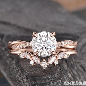 May include: Rose gold engagement ring set with a large, round, clear gemstone and a matching band. The band features a twisted design with small accent stones. The lower band has a marquise and round stone design. The text "SzekiStudio Since 2017" is visible.