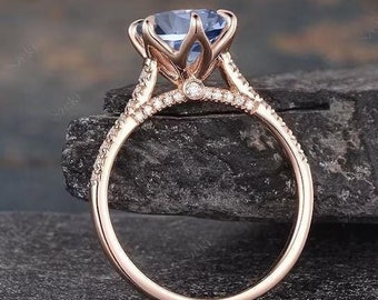 May include: Rose gold engagement ring with a large, light blue gemstone. The band is adorned with small, sparkling stones. The ring is set against a dark, textured background, showcasing its intricate design and elegant style.