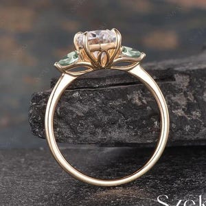 Op de afbeelding: Een gouden verlovingsring met een grote, ronde, heldere middelste steen en twee kleinere, ovale, groene stenen aan weerszijden. De ring ligt op een donkergrijze achtergrond.