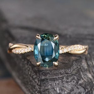 以下が含まれることがあります： ねじれたバンドと大きな楕円形のターコイズブルーのサファイアをセットしたゴールドの婚約指輪。バンドは小さなダイヤモンドで飾られています。