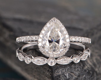 May include: A pear-shaped diamond engagement ring with a halo setting and a matching wedding band. The ring is set in silver and features a delicate design.