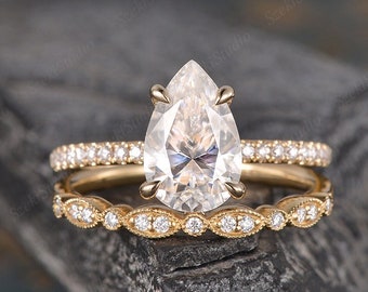May include: A pear-shaped diamond engagement ring set in yellow gold with a matching wedding band. The engagement ring features a large pear-shaped diamond with smaller diamonds set in a pave setting around the band. The wedding band is a delicate band with smaller diamonds set in a pave setting.