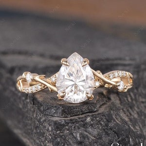 May include: A pear-shaped diamond engagement ring with a twisted band set with smaller diamonds. The ring is made of gold and is set on a dark gray background.
