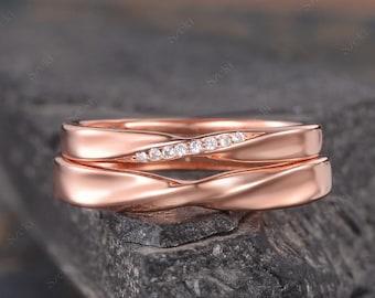 May include: Two rose gold wedding rings. The top ring features a row of small, clear gemstones. The rings have a twisted design. The rings are displayed on a dark, textured surface.