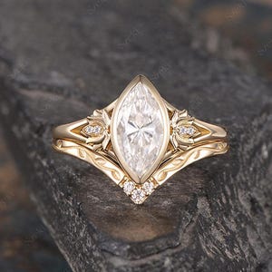 May include: A marquise-cut diamond engagement ring with a gold band, set with small accent diamonds. The ring is on a dark gray stone surface. The band features floral and leaf designs.