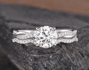 May include: A silver engagement ring set with a round center stone and a twisted band with smaller stones. The ring is on a dark gray background.