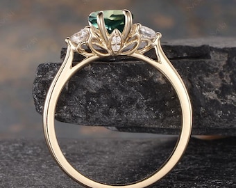 Op de afbeelding: Een gouden ring met een grote, lichtgroene edelsteen in het midden, geflankeerd door twee kleinere, heldere edelstenen. De ring heeft een delicaat, bloemgeïnspireerd ontwerp met kleine accentstenen. De ring wordt gepresenteerd op een donkere, getextureerde ondergrond.
