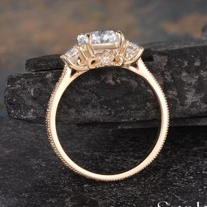 May include: A gold engagement ring with a large, clear center stone and smaller side stones. The ring has a detailed, textured band and is set against a dark, textured background. The word "Szeki" is visible.