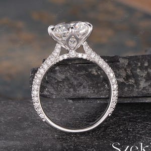 May include: A close-up of a diamond ring with a large, clear center stone. The ring has a pavé band and a detailed setting. The ring is set against a dark, textured background. The word "Szeki" is at the bottom.