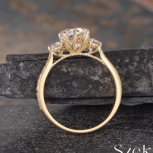 May include: A gold engagement ring featuring a large, clear central gemstone and two smaller side stones. The ring has an intricate, decorative setting and a slender band. The ring is set against a dark, textured backdrop.