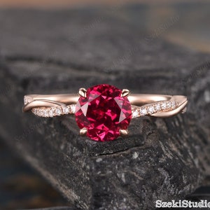 May include: A rose gold engagement ring with a twisted band and a round red ruby gemstone. The band is set with small diamonds.