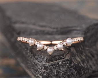 May include: Rose gold ring with a curved design, featuring small pearls and sparkling accent stones. The ring is set on a dark, textured surface, showcasing its elegant details. The ring is a piece of jewelry.
