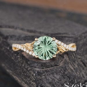 Pode incluir: Um anel de noivado de ouro com uma gema verde e uma banda torcida adornada com pequenos diamantes.