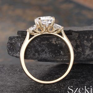 K&ouml;nnte beinhalten: Ein goldener Verlobungsring mit einem gro&szlig;en, rechteckigen Mittelstein und zwei kleineren, trapezf&ouml;rmigen Seitensteinen. Der Ring hat eine filigrane, verzierte Fassung. Das Wort "Szeki" ist sichtbar.