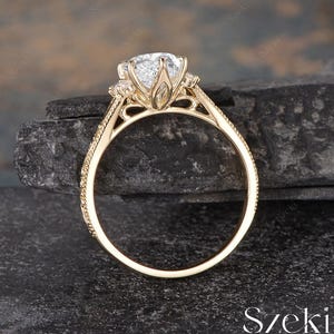 May include: A gold engagement ring with a large, clear gemstone. The ring features a detailed filigree design around the center stone and along the band. The ring is set against a dark, textured background.