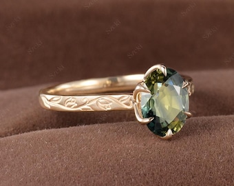 Könnte beinhalten: Ein goldener Ring mit einem ovalen, olivgrünen Edelstein. Das Band des Rings weist ein eingraviertes Blumenmuster auf. Der Edelstein wird von vier Krappen gehalten. Der Ring ist vor einem braunen Hintergrund platziert.