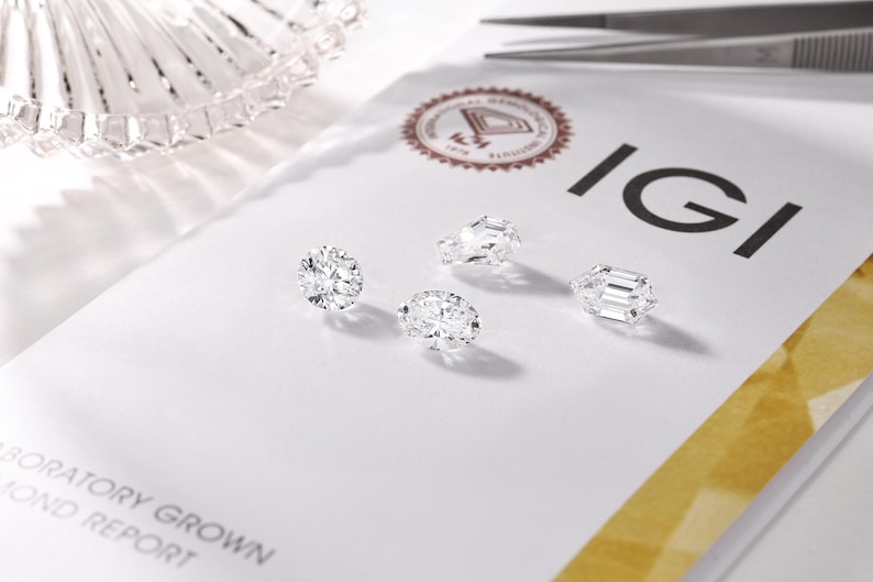 May include: Four sparkling, laboratory-grown diamonds of varying cuts are displayed on a white surface. The IGI logo and text are visible, along with a pair of tweezers and a decorative glass dish.